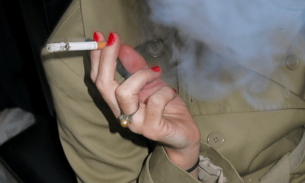 Gros plan dans la nuit avec un flash d'une femme en imperméable kaki, qui tient dans sa main aux ongles rouges une cigarette consumée. De la fumée habille la photo.