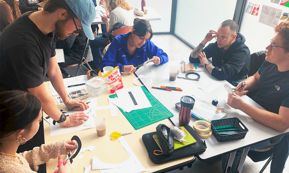 Sur une table d'école, des étudiants découpent et collent des éléments sur des grilles graphiques.